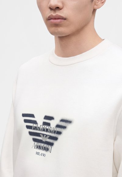 Jeune personne portant un sweat blanc avec un logo Emporio Armani noir et blanc et le mot Milano sur la poitrine.