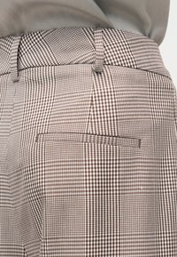 Pantalon à carreaux marron et blanc avec une poche passepoilée à l'arrière et des passants pour ceinture, porté avec un haut vert olive rentré à l'intérieur.
