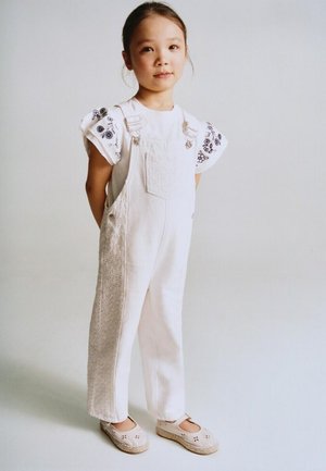 Jeune fille debout, les mains derrière le dos, portant une salopette blanche par-dessus une chemise à manches courtes à motif floral et des chaussures beige.