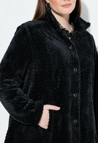Svart plyschkappa med ett texturerat diamantmönster, försedd med krage, knappstängning och sidofickor. Slät tyg och mjuk utseende.
