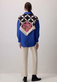 Donna che indossa una camicia blu a trama con una sciarpa nera, bianca e con motivo floreale con frange sul retro, pantaloni color crema e stivali neri.