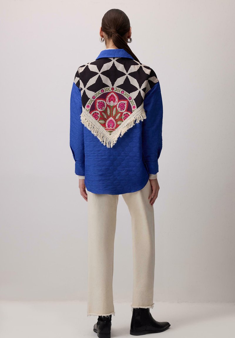 Donna che indossa una camicia blu a trama con una sciarpa nera, bianca e con motivo floreale con frange sul retro, pantaloni color crema e stivali neri.