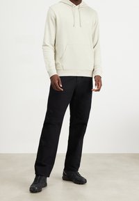 Ljusbeige hoodie med framficka och dragsko i huvan, kombinerad med svarta byxor och svarta sportskor. Diskret logotyp på bröstet.