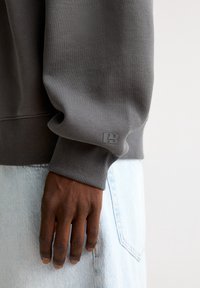 Szara bluza z ribbowanymi mankietami i subtelnym logo. Gładka tekstura, luźny krój, w połączeniu z jasnoniebieskimi dżinsami, ukazująca sposób trzymania rąk.
