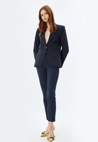 Blazer blu navy a righe con chiusura a un bottone, abbinato a pantaloni blu aderenti e décolleté dorati a punta lucida. Design semplice e su misura.