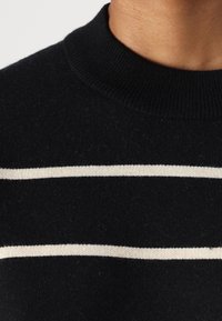 Gros plan sur un pull en maille noire avec deux rayures blanches horizontales et un col rond côtelé porté par une personne.