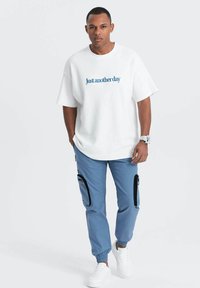 T-shirt oversize blanche avec le texte bleu "Just another day", associé à un pantalon de jogging bleu clair avec des poches zippées noires, et des baskets blanches.