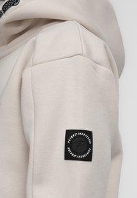 Beige hoodie met een gladde textuur, voorzien van een zwart logo-opdruk op de linker mouw en een trekkoordkap. Eenvoudig ontwerp en casual pasvorm.