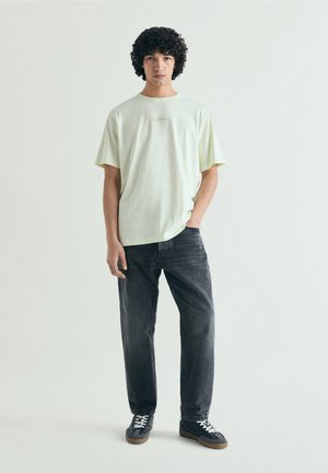 Joven con pelo rizado está de pie con una camiseta verde pálido, vaqueros negros y zapatillas negras frente a un fondo liso y claro.