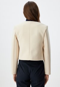 Lyhennetty kerma blazer, jossa on muotoillut hartiat, sileä pinta ja suljettu takasuunnittelu. Yhdistetty tummiin housuihin, korostaen hankkeen istuvuutta.