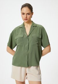 Kurzärmliges grünes Shirt mit strukturiertem Finish, mit einer Knopfleiste, zwei Brusttaschen und lockerem Schnitt. Kombiniert mit hellen Shorts.