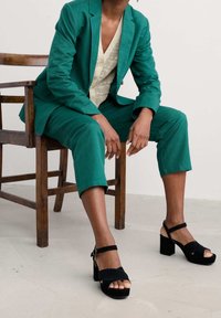 Blazer teal e pantaloni cropped in tessuto texturizzato, abbinati a sandali con piattaforma nera caratterizzati da una fascia larga e una punta aperta.