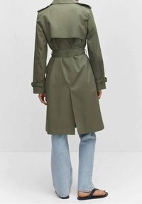 Olivgrüner Trenchcoat mit tailliertem Gürtel, Rückeneinsatz und krempelbaren Ärmeln, kombiniert mit hellblauen, weit geschnittenen Jeans und schwarzen Sandalen.