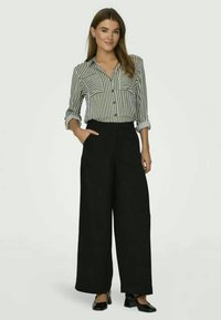 Kvinne står med hånden i lommen, iført svarte vide bukser, svarte hæler og en svart-hvitt stripete skjorte med knapper.