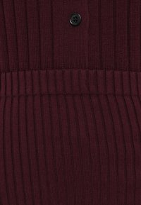 Burgundy kötött pulóver függőleges bordás textúrával, kerek nyakkivágással és egy fekete gombbal az elején.