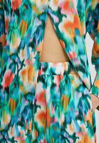 Chemise à imprimé abstrait multicolore boutonnée partiellement ouverte, associée à un pantalon assorti à taille élastique, laissant voir le ventre et une main dans la poche.