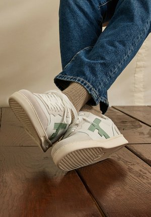 Witte sneakers met een groen logo en beige sokken, gedragen met blauwe spijkerbroek, rustend op een donkere houten vloer.
