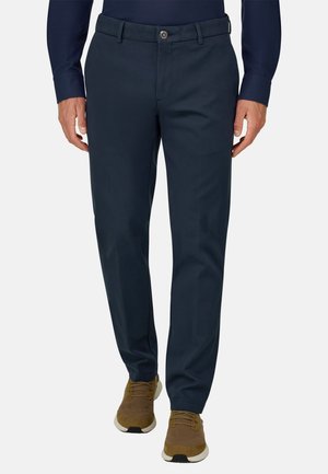 Homme portant un pantalon slim bleu marine, une chemise à manches longues bleu foncé et des baskets marron, debout devant un fond blanc.