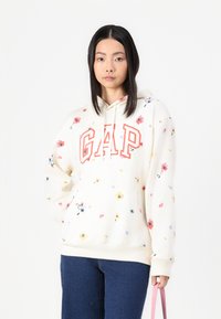 Sudadera blanca con estampado floral en tonos rosa y amarillo. Presenta un bolsillo frontal y el logo "GAP" en letras rojas. El modelo lleva jeans azules.