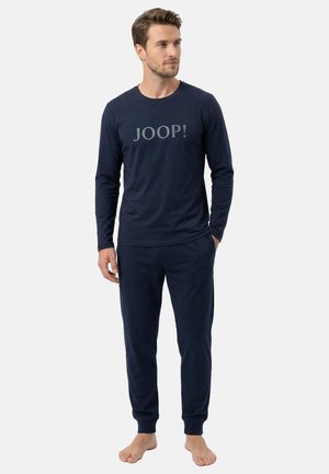 Mann steht barfuß und trägt ein marineblaues Langarmshirt mit dem Schriftzug „JOOP!“ und passende marineblaue Freizeithosen vor einem einfarbigen Hintergrund.