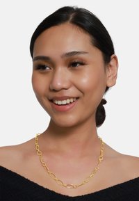 Gold Necklace mit ovalen Gliedern, texturierten Details und einem sicheren Verschluss, getragen auf einem nackten Schlüsselbein vor einem einfarbigen weißen Hintergrund.