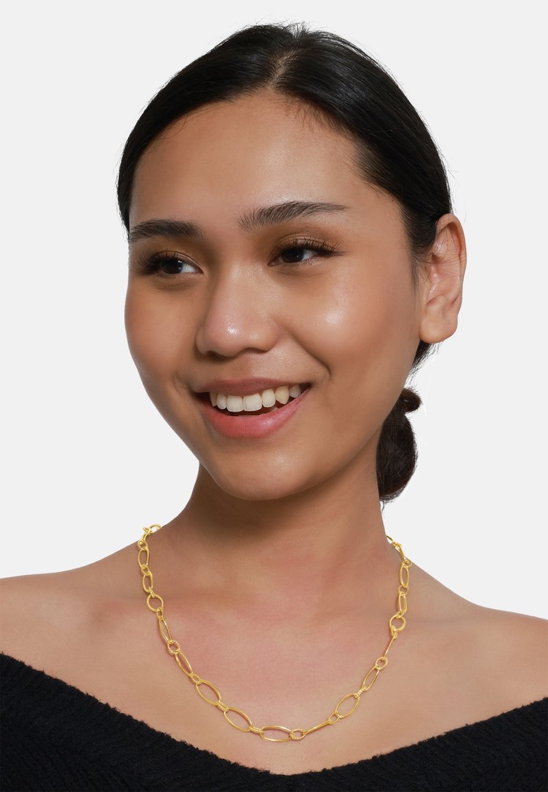 Gold Necklace mit ovalen Gliedern, texturierten Details und einem sicheren Verschluss, getragen auf einem nackten Schlüsselbein vor einem einfarbigen weißen Hintergrund.