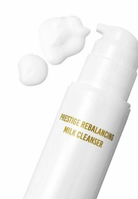 Biała plastikowa butelka z pompką oznaczona "Prestige Rebalancing Milk Cleanser" w kolorze złotym. Obok butelki znajdują się dwie małe krople kremowego środka czyszczącego.
