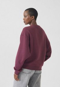 Felpa oversize color marrone con scollo tondo e spalle cadenti, abbinata a jeans grigi chiari. Texture liscia con polsini a costine.