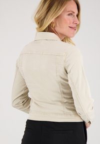 Vrouw met blond haar, gekleed in een crèmekleurige jas en gouden ringoorbellen, zijwaarts gedraaid met een lichte glimlach, gekleed in zwarte broek.