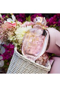 Bouteille de parfum Juicy Couture Just Moi dans un panier en osier blanc avec des fleurs roses et blanches, sur un fond de fleurs colorées.