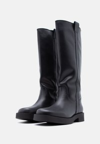 Μαύρα δερμάτινα μπότες μέχρι το γόνατο με στρογγυλεμένη μύτη, επίπεδη σόλα, ελάχιστες ραφές και μικρές θηλιές στην κορυφή για εύκολη τοποθέτηση.