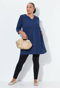 Donna in tunica blu con motivo, leggings neri, ballerine beige, che tiene in mano una borsa beige con texture, in piedi di fronte a uno sfondo bianco.
