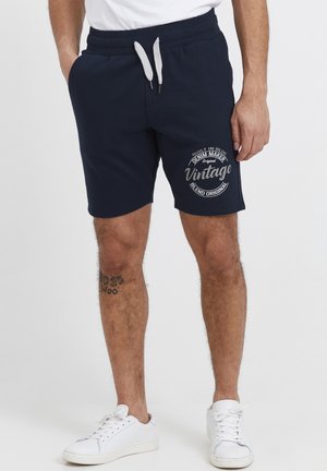 Shorts en coton bleu marine avec une taille élastique et un cordon de serrage gris. Présente un design graphique textuel circulaire blanc sur la cuisse gauche.