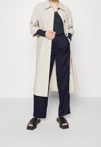 Beige lightweight trench coat with button closure, black long-sleeve top, navy wide-leg pants, and black platform sandals.