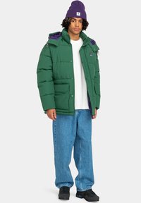 Chaqueta acolchada verde con detalles morados y bolsillos con cremallera, combinada con jeans de mezclilla holgados y un gorro morado. Tejido texturizado, diseño casual.