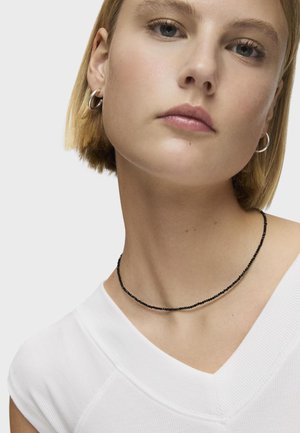 Collana choker di perle nere indossata su un collo di carnagione chiara, che completa una maglietta bianca a V. Design semplice e delicato con piccole perle rotonde.