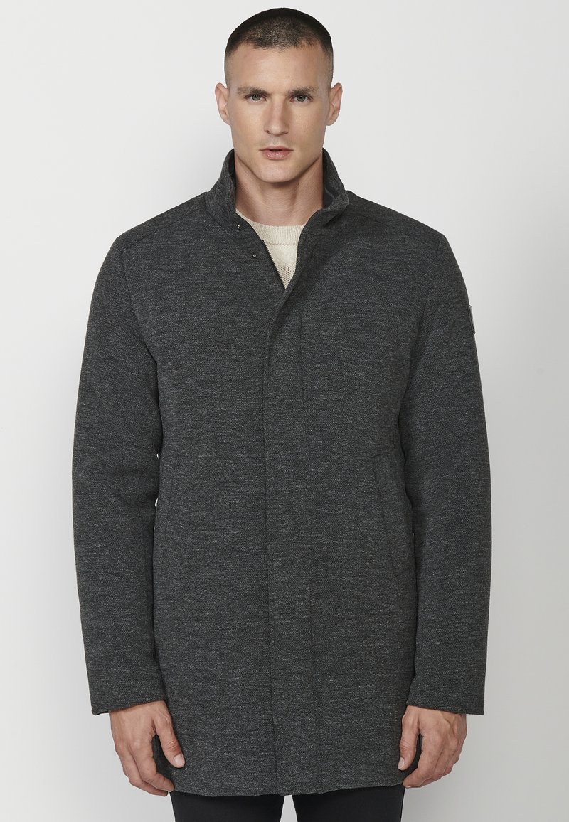 Abrigo gris oscuro con un cuello alto, hecho de una tela texturizada. Presenta un cierre frontal con botones a presión y bolsillos laterales. Diseño simple y aerodinámico.