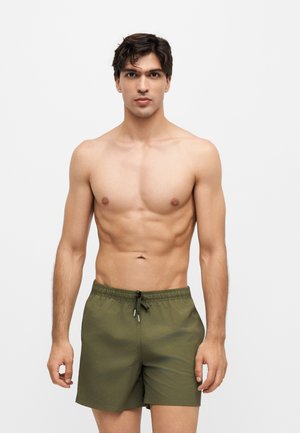 Ung mand med kort mørkt hår iført olivengrøn badeshorts, der står mod en ensfarvet lys baggrund.