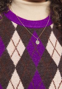 Personne portant un col roulé beige sous un pull à losanges violet, marron et crème, avec un collier pendentif en or.