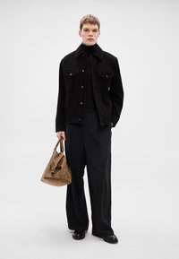 Melnā kordurojas jaka ar divām krūšu kabatām, pār kuras uzvilkts melns augsts apkakles džemperis, kombinēta ar platām melnām biksēm un brūnu somu.