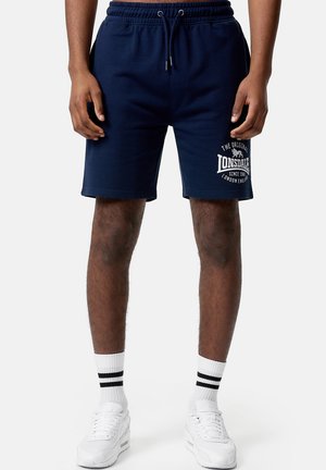 Shorts en coton bleu marine avec une taille élastique, poches avant et impression du logo blanc sur la jambe gauche. Associés à des chaussures de sport blanches et des chaussettes rayées.