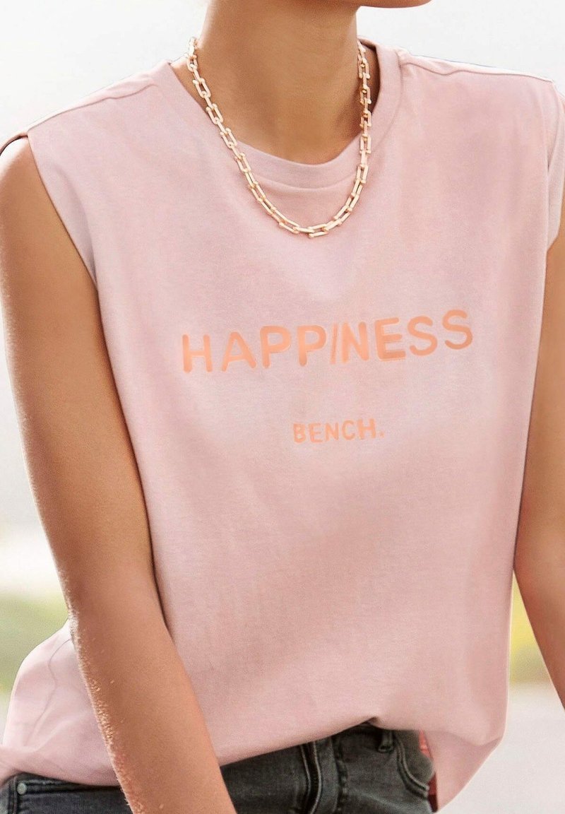 Chemise rose sans manches avec le texte « HAPPINESS BENCH. », portée avec un collier épais en chaîne dorée et un jean foncé.