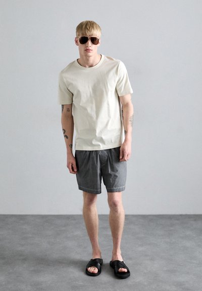 Tricou gri deschis, pantaloni scurți gri închis și sandale negre. Persoana poartă ochelari de soare; design minimalistic cu mâneci scurte și croială lejeră.