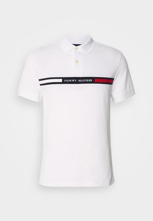 Bijela polo majica s klasičnim ovratnikom, kratkim rukavima i horizontalnim pojasom s mornarsko plavim i crvenim prugama s natpisom "TOMMY HILFIGER" u sredini.