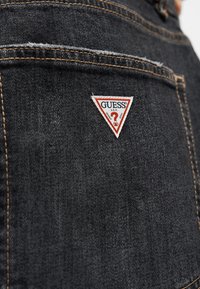 Το σκούρο τζιν ύφασμα διαθέτει ένα τριγωνικό κόκκινο και λευκό λογότυπο με το "GUESS" και ένα ερωτηματικό, διακοσμημένο με αντίθετη χρυσή ραφή.