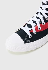 Fekete vászon tornacipő fehér gumi talppal és orrmerevítővel, piros kiemelő csíkkal és fehér fűzőkkel, látható fűzőlyukakkal és texturált felülettel.