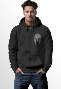 Schwarze Zip-Hoodie mit einer Fronttasche und einem rissigen Spartanerhelm-Graphic in Grau auf der linken Brustseite. Glatte Textur, lässige Passform.