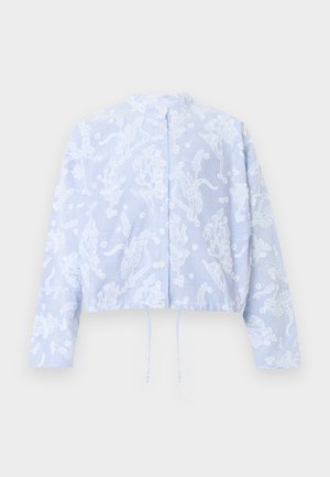 Nő áll, világoskék virágmintás laza dzsekit és széles szárú nadrágot visel, fehér felsővel és bordó balerinával párosítva.