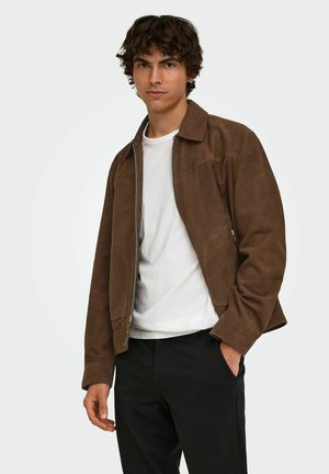 Jeune homme portant une veste marron zippée sur une chemise blanche et un pantalon noir, debout avec une main dans la poche sur un fond uni.