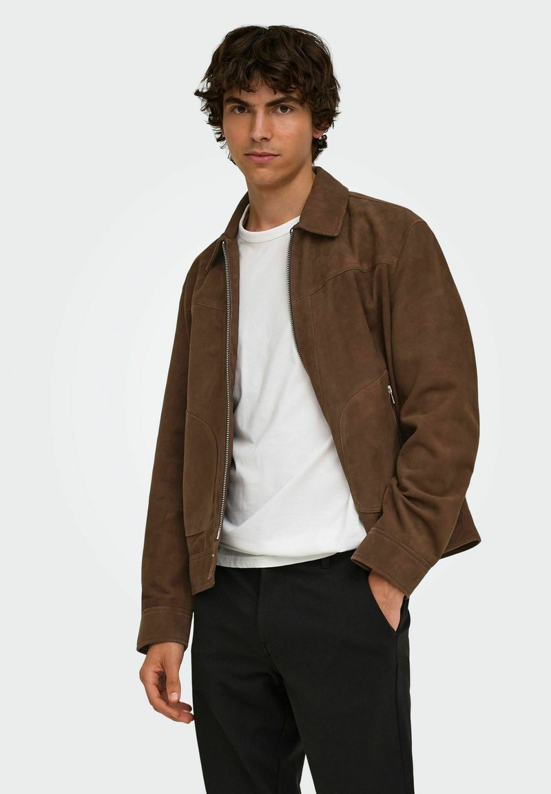 Jeune homme portant une veste marron zippée sur une chemise blanche et un pantalon noir, debout avec une main dans la poche sur un fond uni.